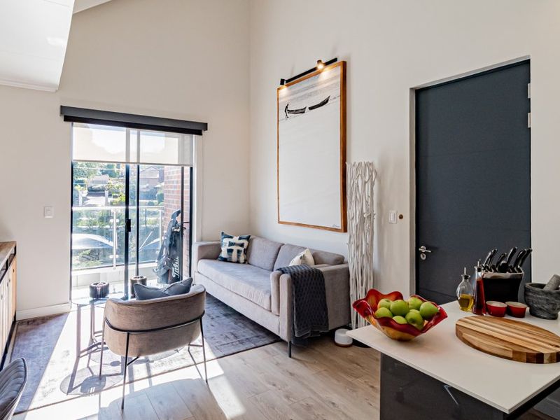 Bright open-plan living area featuring vaulted ceilings, modern laminate flooring, and direct access to a private balcony through sliding glass doors.