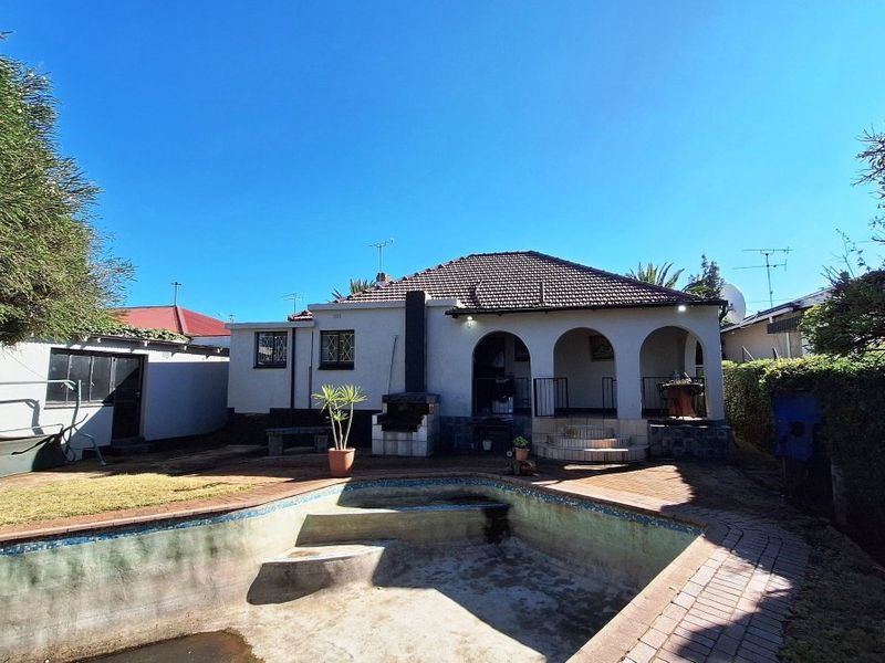 Rear view of the property featuring a covered patio, built-in braai area, and a swimming pool requiring maintenance.