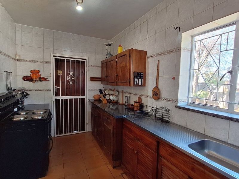 A functional kitchen featuring wooden cabinetry, stone countertops, a stainless steel sink, and tiled finishes, with a security gate and window bars.