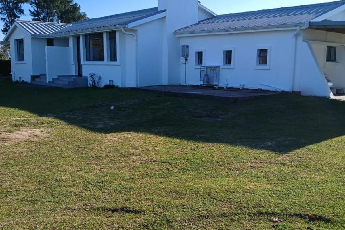 Rear view of a white farmhouse featuring a metal roof, central chimney, and a paved patio area overlooking the surrounding lawn.