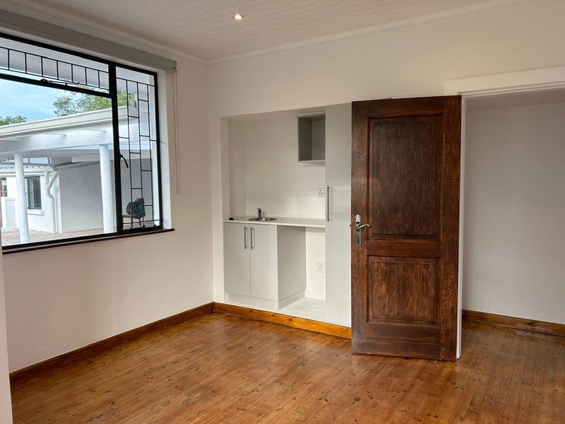 A bright bedroom featuring polished timber flooring, a convenient built-in kitchenette with a sink, and a solid timber door.