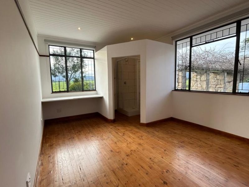 A bright bedroom featuring polished timber flooring, a built-in desk, and an en-suite shower cubicle with large windows overlooking the farm landscape.