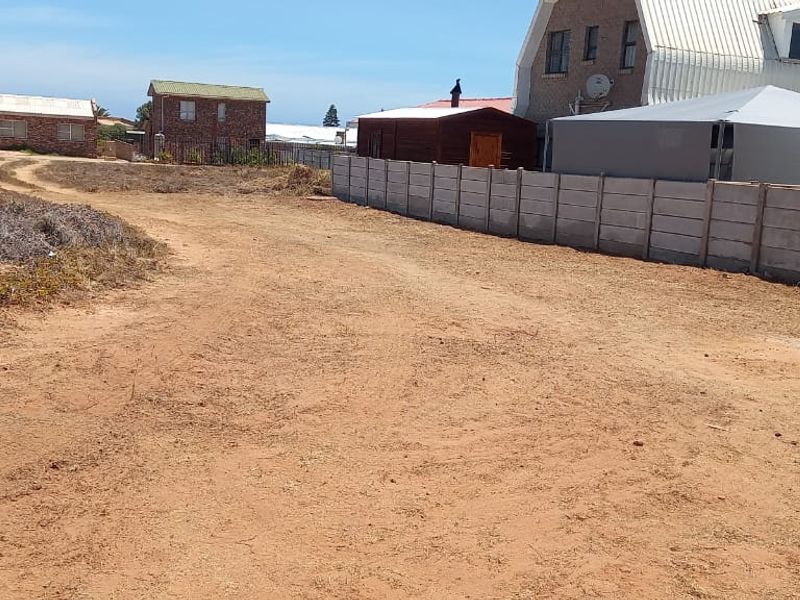 A vacant residential plot featuring an unpaved access track and a precast concrete boundary wall, situated in a developed coastal neighborhood.
