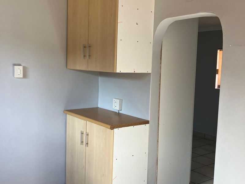A small kitchen area featuring built-in cabinetry with laminate countertops, tiled flooring, and a distinctive timber ceiling, leading through an arched doorway.