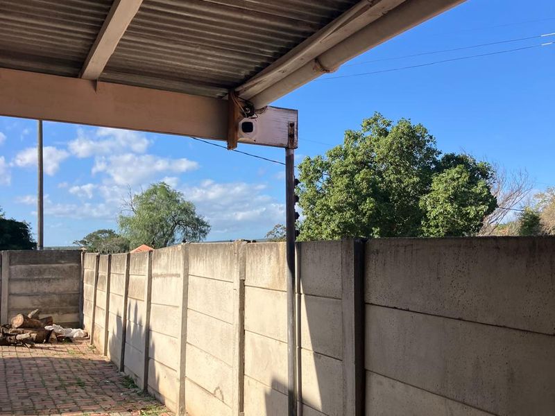 A covered outdoor patio area featuring a corrugated metal roof, paved flooring, and a precast concrete boundary wall with electric fencing.