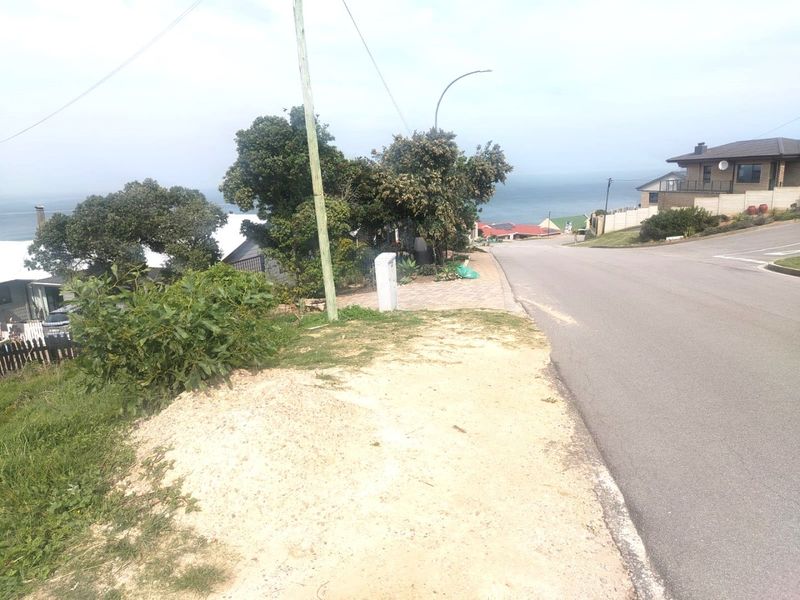 A street-level view of the residential property frontage, showing the paved road, sidewalk, and utility infrastructure with a distant ocean view.