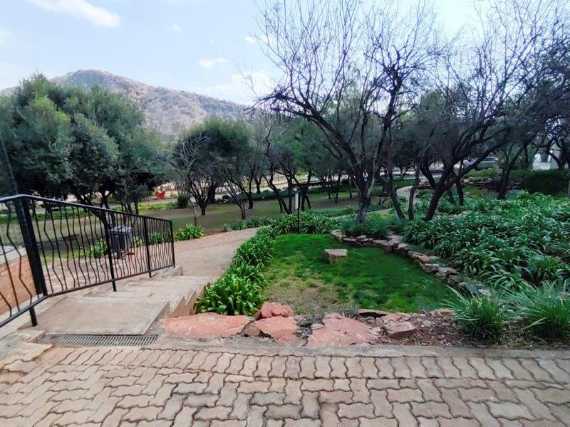 A landscaped garden area featuring a paved walkway, stone retaining walls, and concrete stairs leading to a scenic outdoor space with mature trees.