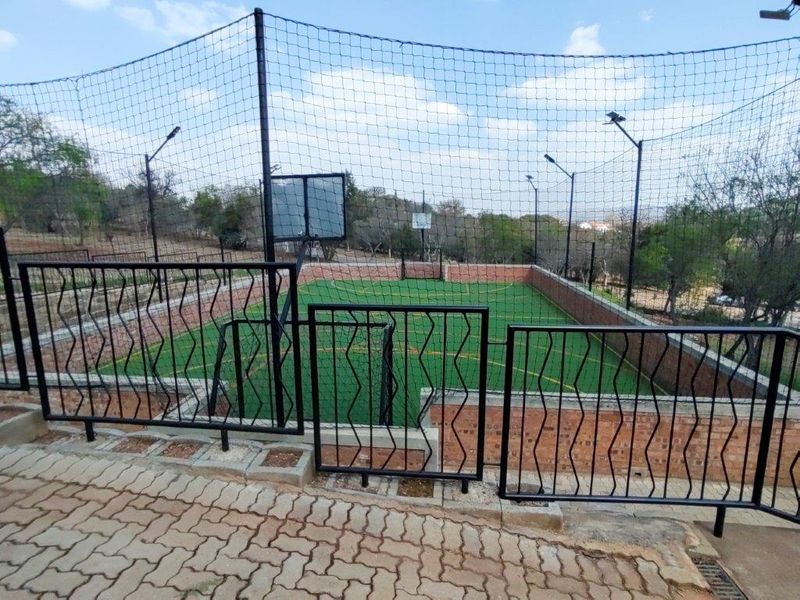 A well-maintained outdoor sports facility featuring an artificial turf court, perimeter safety netting, floodlights, and a basketball hoop, enclosed by brick walls and metal railings.