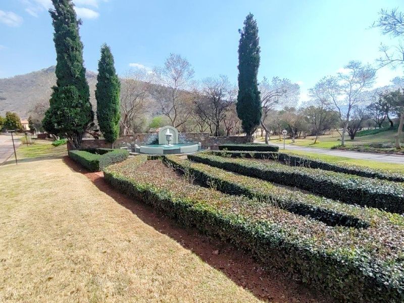 A well-maintained garden area featuring manicured hedges, tall cypress trees, and a central stone fountain with a stone retaining wall.