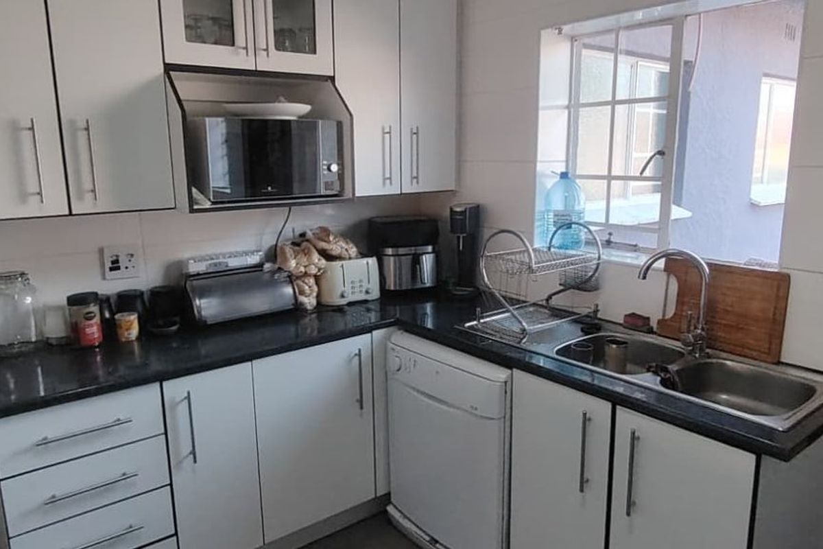 A functional kitchen featuring white cabinetry, dark laminate countertops, a double bowl sink, and tiled splashbacks with a window overlooking the exterior.