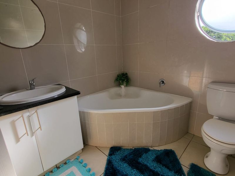 A functional bathroom featuring a corner bathtub, vanity with basin, toilet, and a distinctive porthole window with neutral wall and floor tiling.