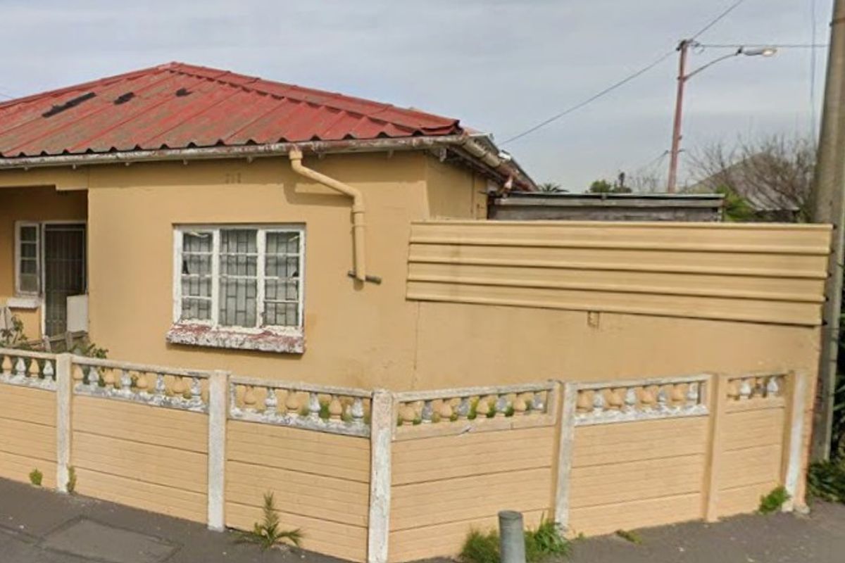 Front elevation of a single-story house featuring a corrugated metal roof, security-barred windows, and a precast concrete boundary wall.