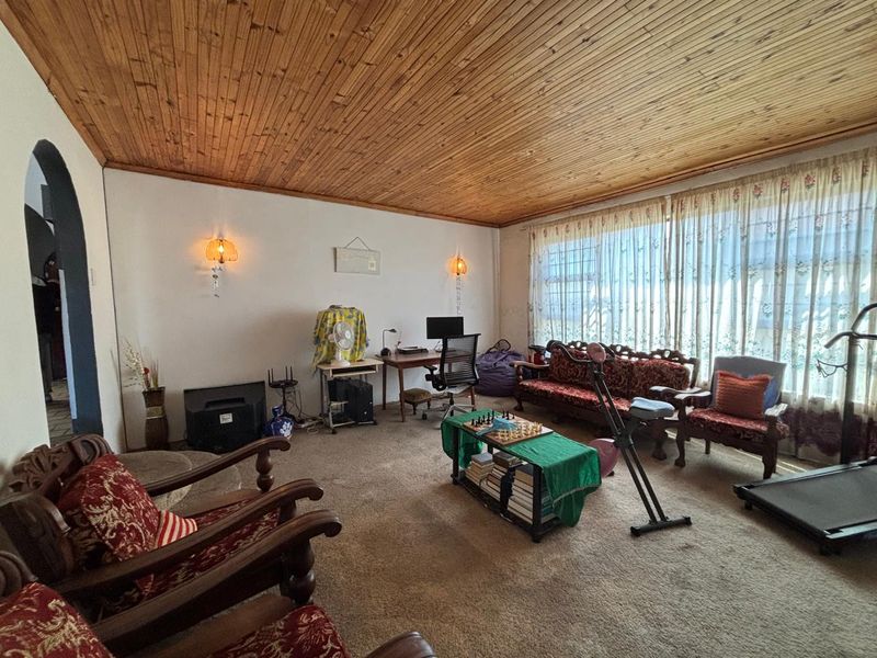 Spacious living room featuring a distinctive timber-clad ceiling, wall-mounted lighting, and large windows, offering a dated but functional space ready for modernization.