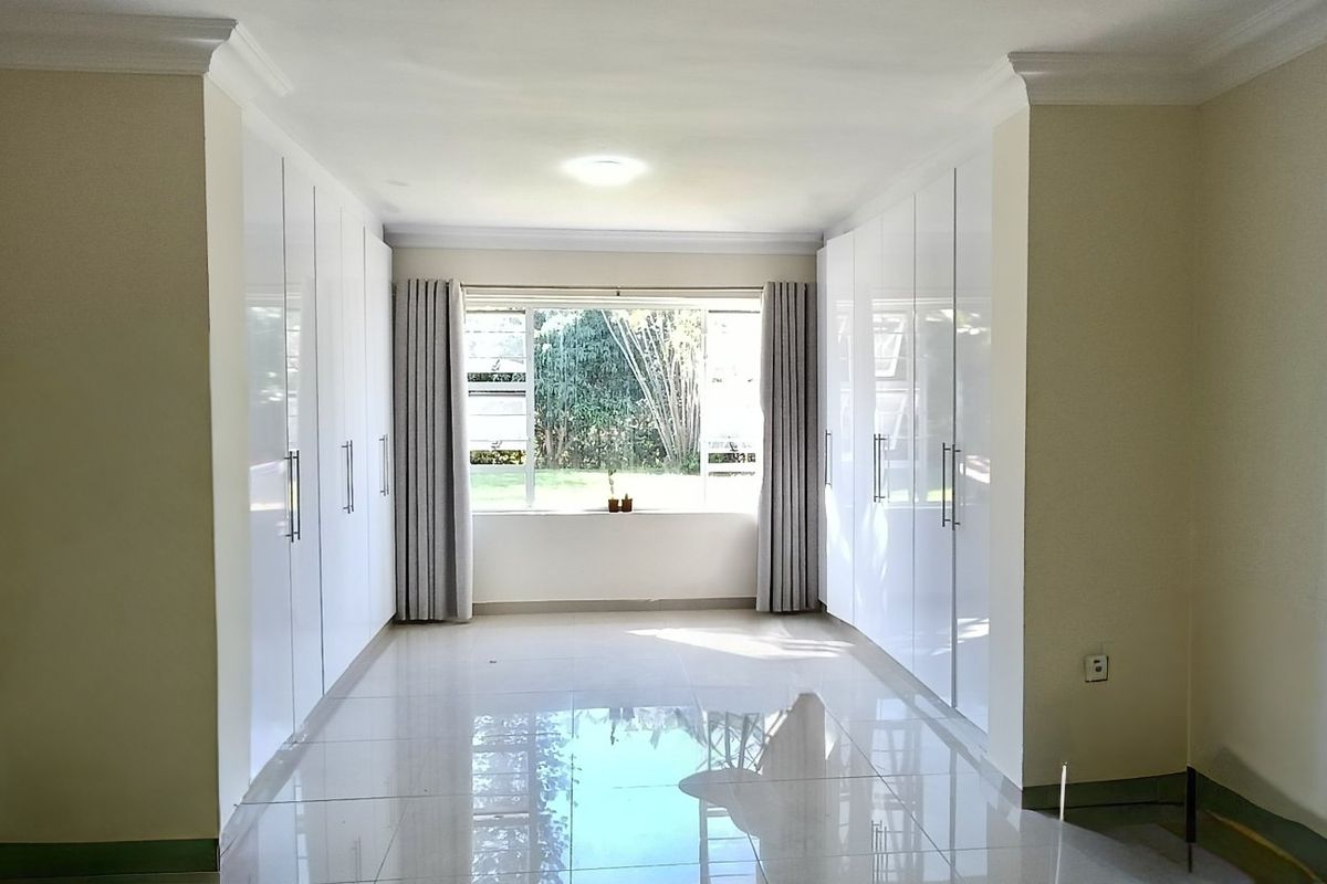 A spacious, empty bedroom featuring extensive white built-in cupboards, polished tiled flooring, and large windows overlooking the garden.