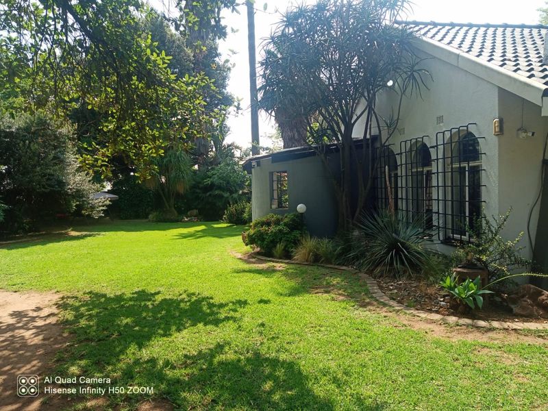 A view of the rear garden featuring a well-maintained lawn, mature trees, and the side elevation of the house with arched windows and security bars.