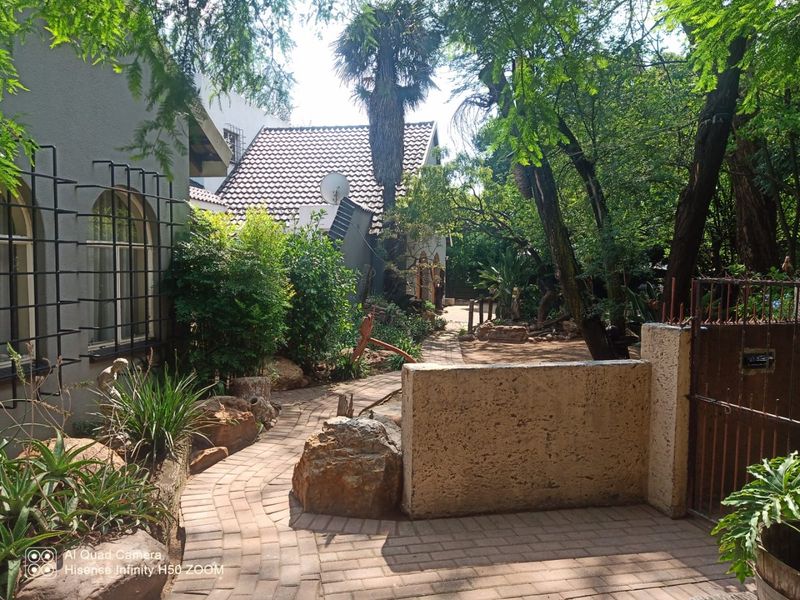 A lush, established garden featuring a winding paved walkway, stone retaining walls, and mature trees surrounding the property exterior.