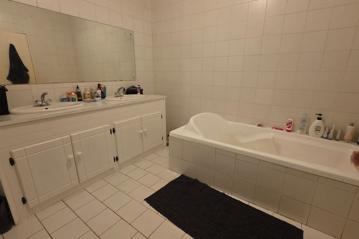 A dated bathroom featuring a double vanity, built-in bathtub, and full-width wall mirror, finished with white ceramic wall and floor tiling.
