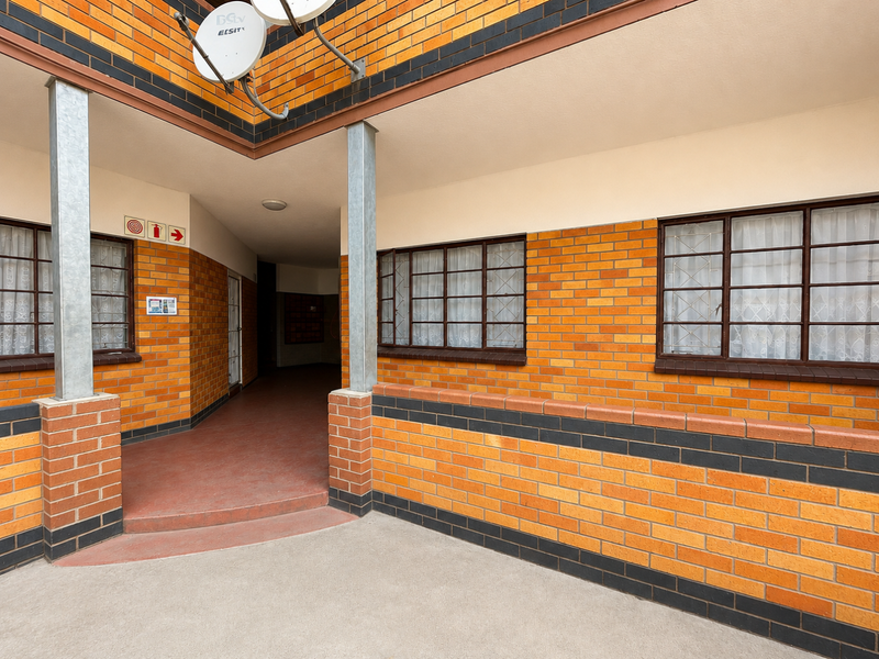 Exterior view of an apartment building entrance featuring face brick walls, steel-framed windows with security bars, and a paved walkway.