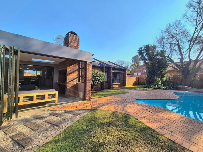 Rear garden featuring a swimming pool, brick paved patio, and a covered outdoor entertainment area with a built-in braai and folding glass doors.
