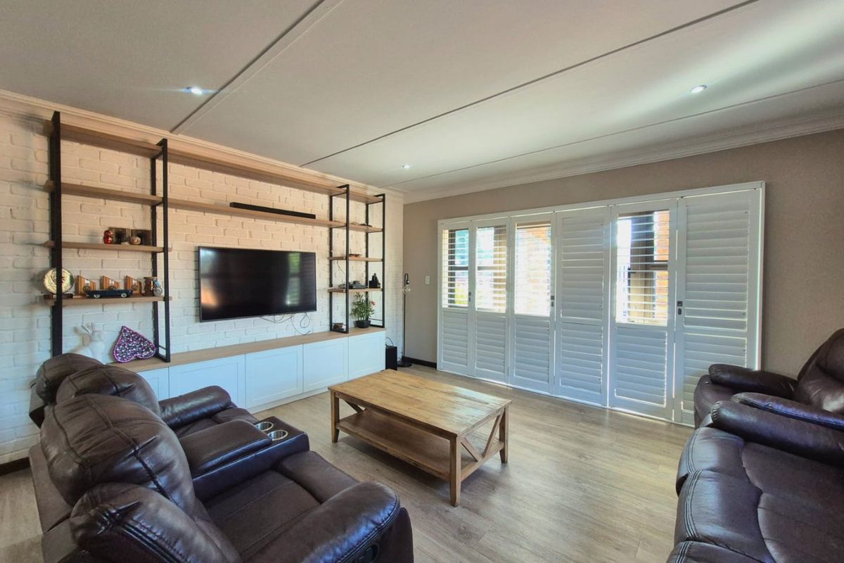 A well-maintained living room featuring a white painted brick feature wall with built-in shelving, laminate flooring, and plantation shutters opening to the exterior.