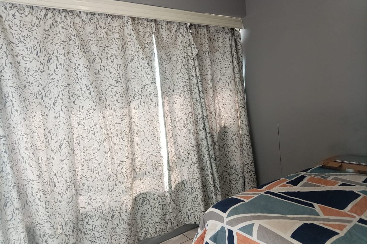 A bedroom featuring tiled flooring and a large window covered by curtains, showing a visible vertical crack on the wall.