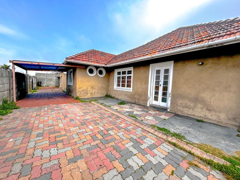 Front exterior view of a single-story house featuring a tiled roof, paved driveway, carport, and distinctive porthole windows, showing signs of wear.
