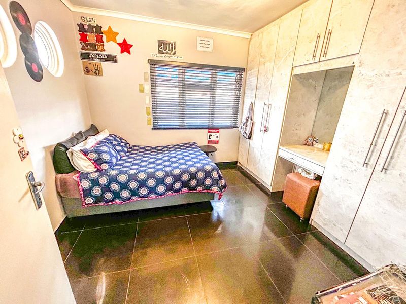 A well-maintained bedroom featuring dark tiled flooring, extensive built-in cupboards with an integrated vanity, and venetian blinds for privacy.