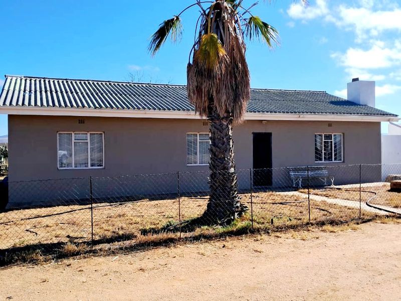 Front elevation of a single-story farm dwelling featuring a corrugated metal roof, chimney, and security-barred windows, set on a dry, fenced plot.