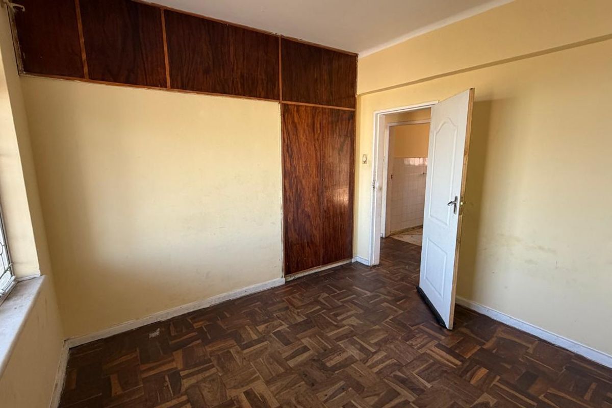 An empty bedroom featuring original parquet flooring, built-in wooden cupboards, and security bars on the window, requiring cosmetic updates to the walls and flooring.