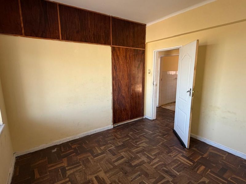 An empty bedroom featuring original parquet flooring, built-in wooden cupboards, and security bars on the window, requiring cosmetic updates to the walls and flooring.