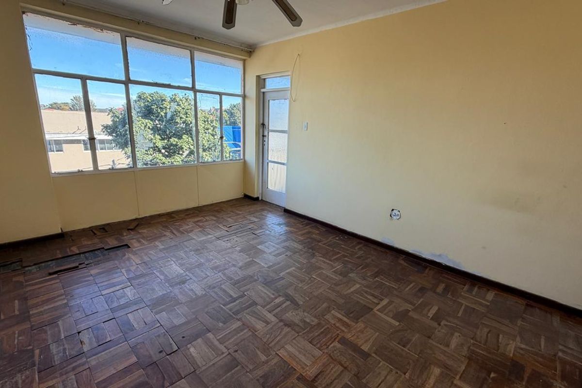 An empty living room featuring original parquet flooring, a ceiling fan, and large windows providing natural light. The space requires renovation and floor repairs.