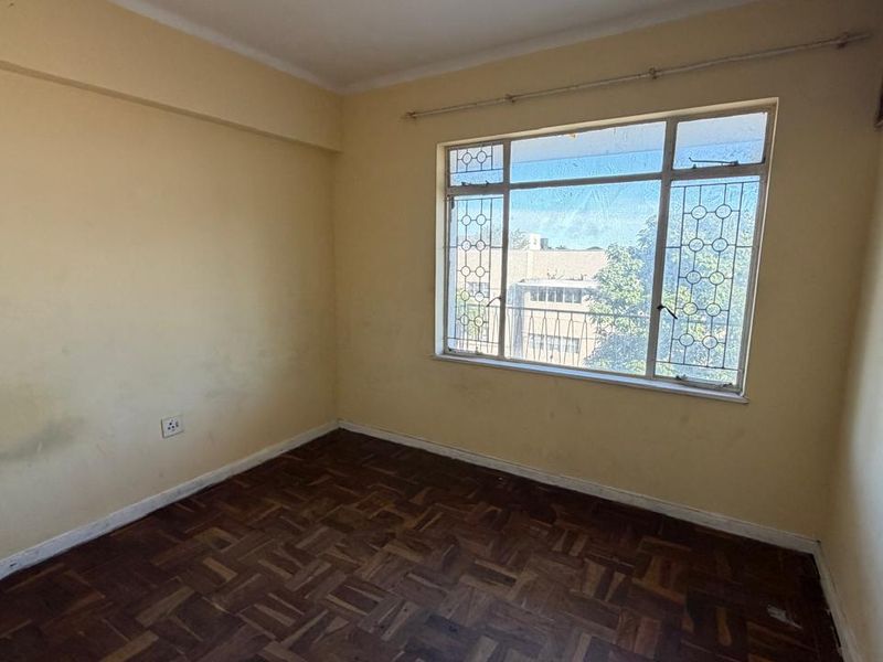 An empty bedroom featuring original parquet flooring, a large window with security bars, and walls requiring cosmetic attention.