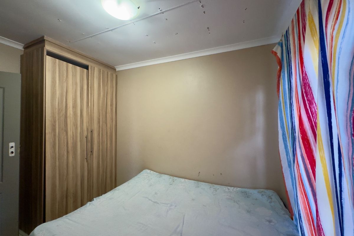 A compact bedroom featuring a built-in wood-grain wardrobe and standard ceiling lighting, requiring minor cosmetic updates to the walls and ceiling.