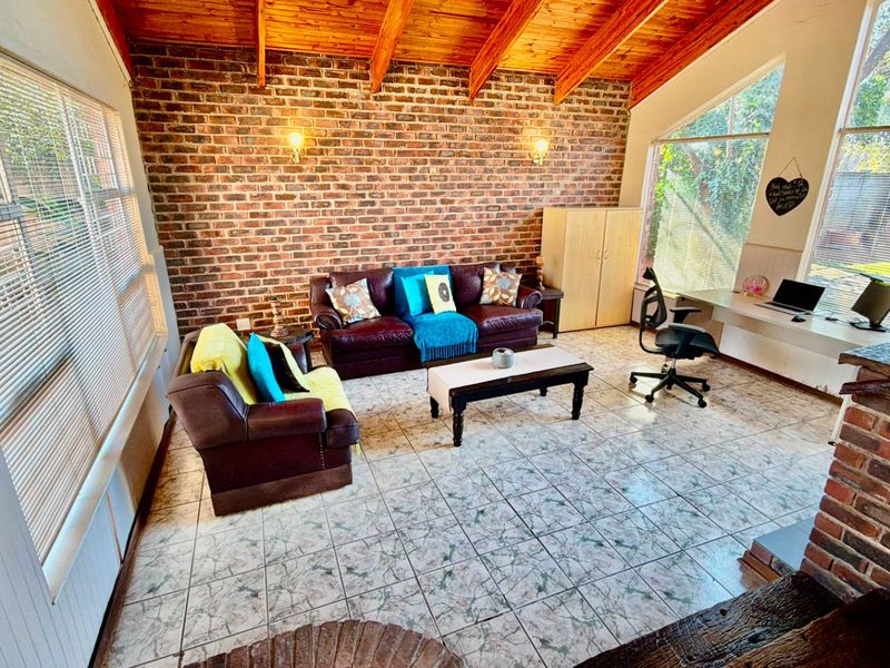 Spacious living area featuring exposed brick walls, timber ceiling beams, tiled flooring, and a built-in desk space with large windows providing natural light.