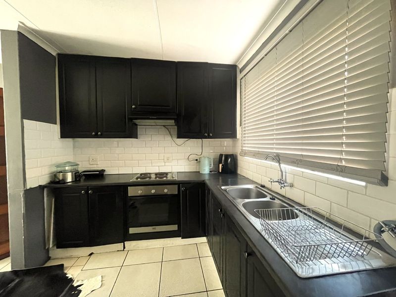 A functional kitchen featuring black cabinetry, subway tile backsplash, built-in oven, electric hob, and a double bowl stainless steel sink with venetian blinds.