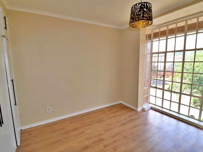 A bright, empty bedroom featuring laminate flooring, built-in cupboards, and a large steel-framed window providing natural light.
