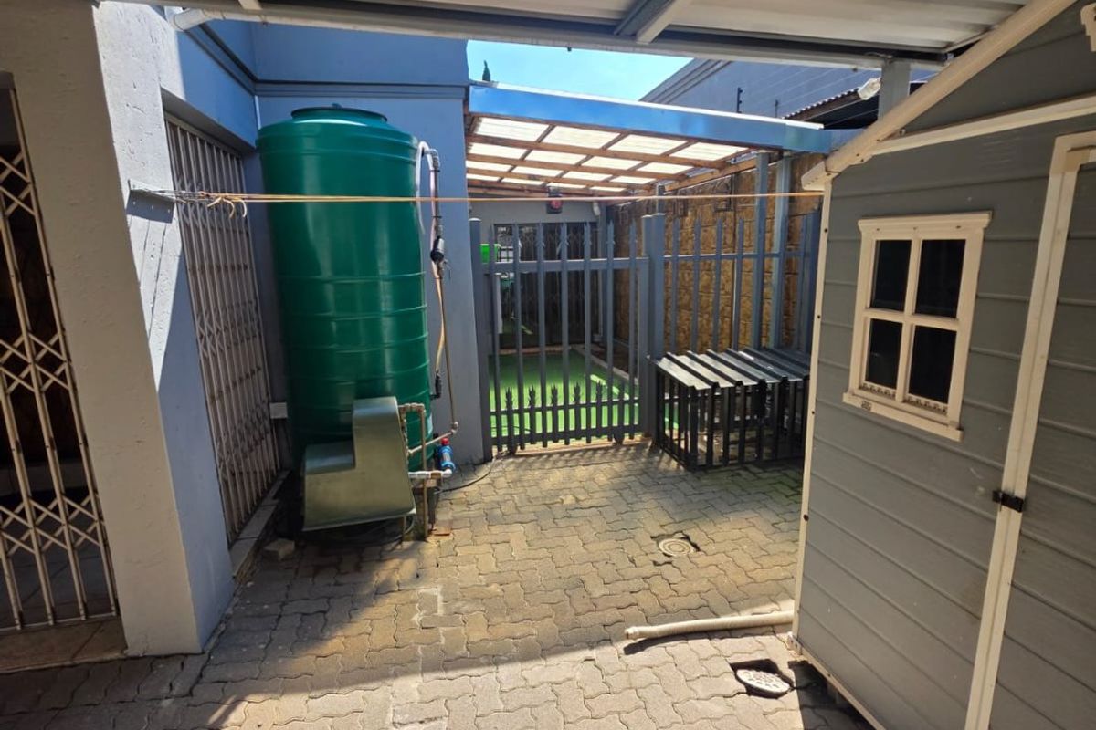 A paved rear courtyard area featuring a water storage tank, a metal carport structure, and a small storage shed with security gates.