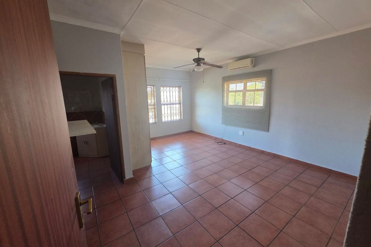 A spacious, empty living area featuring tiled flooring, a ceiling fan, and a wall-mounted air conditioning unit, with an adjacent room visible.