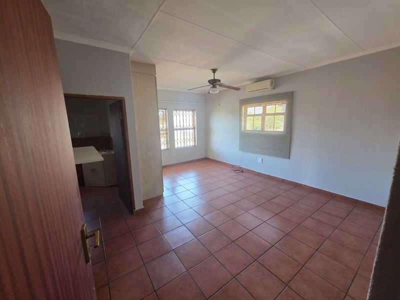 A spacious, empty living area featuring tiled flooring, a ceiling fan, and a wall-mounted air conditioning unit, with an adjacent room visible.