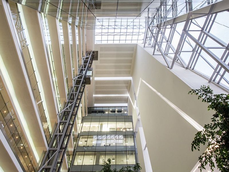 A modern commercial atrium featuring a soaring glass skylight, multi-level mezzanine walkways, and a prominent glass elevator shaft.