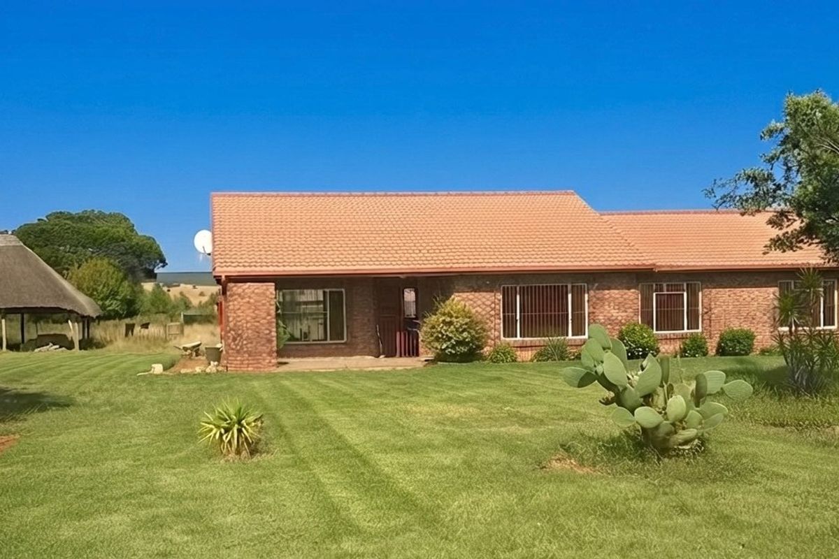 A single-story face brick farmhouse featuring a tiled roof, security bars on windows, and a separate thatched lapa set within a spacious garden.