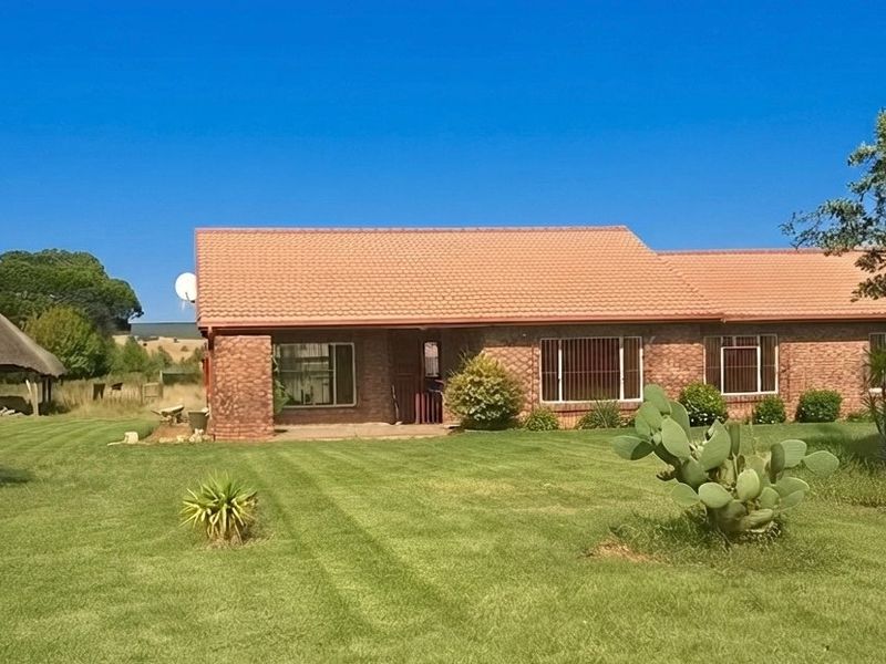A single-story face brick farmhouse featuring a tiled roof, security bars on windows, and a separate thatched lapa set within a spacious garden.