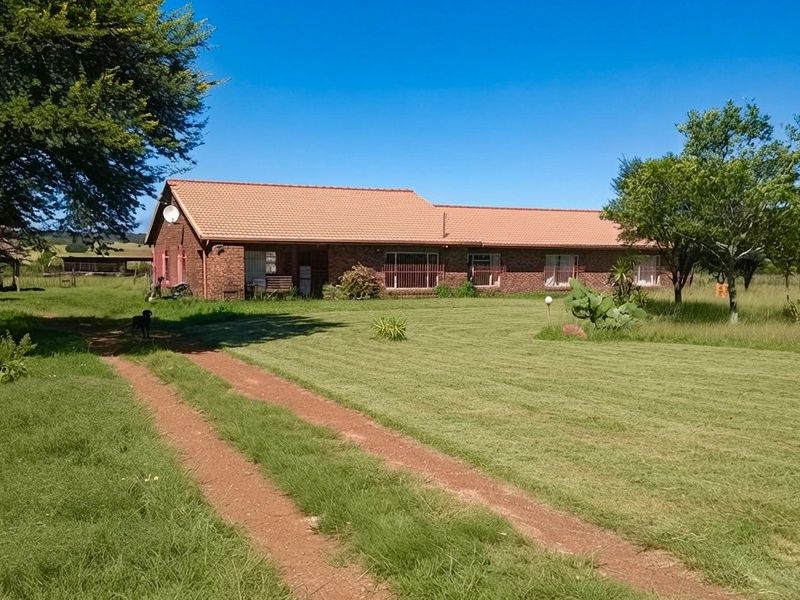 A single-story brick farmhouse with a tiled roof, set on a large grassy plot with a dirt driveway approach.