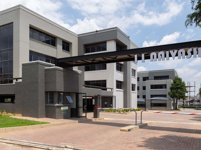 Modern commercial office complex entrance featuring a secure gatehouse, paved driveway, and contemporary multi-story building architecture with extensive glass facade elements.