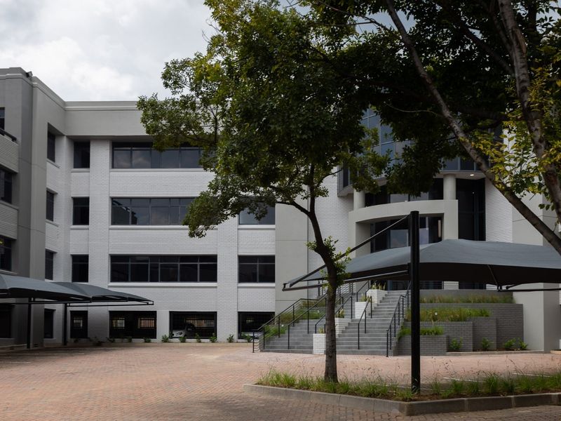 A professional view of a multi-story commercial building featuring a paved parking area, shade cloth carports, and a prominent entrance staircase.