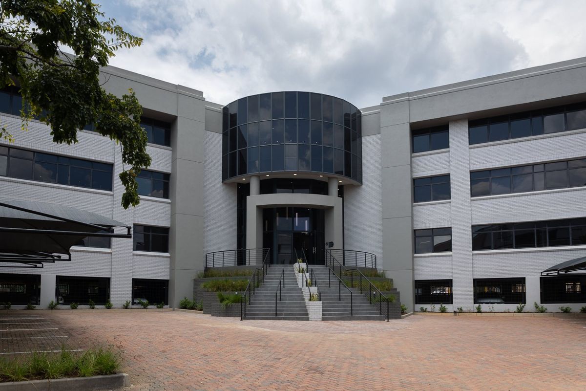 Modern commercial office building featuring a prominent glass facade, central staircase entrance, and paved driveway with covered parking.
