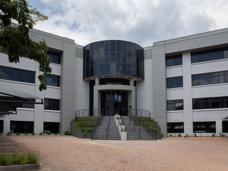 Modern commercial office building featuring a prominent glass facade, central staircase entrance, and paved driveway with covered parking.