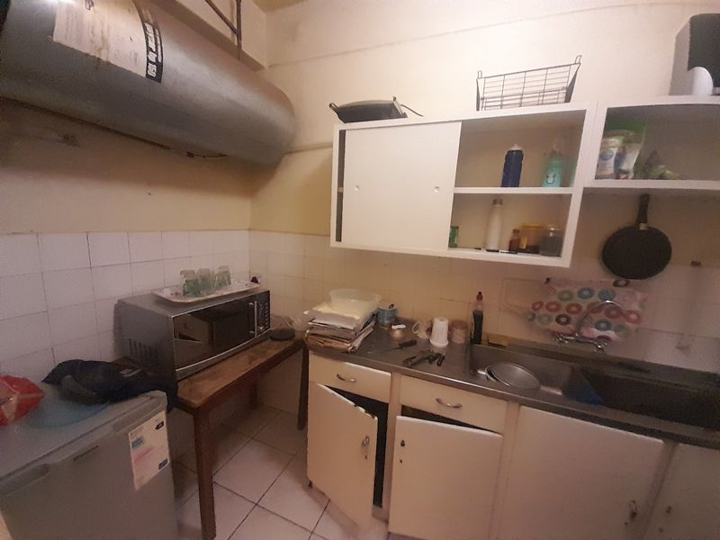 A compact kitchen area featuring a stainless steel sink, wall-mounted geyser, and dated cabinetry requiring maintenance or renovation.