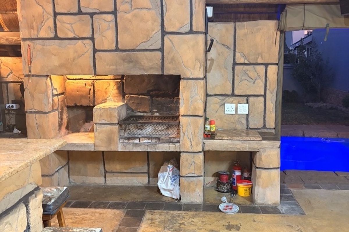 Covered outdoor patio featuring a large stone-clad built-in braai, concrete prep surfaces, and a thatched roof, overlooking a swimming pool.