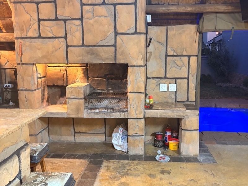 Covered outdoor patio featuring a large stone-clad built-in braai, concrete prep surfaces, and a thatched roof, overlooking a swimming pool.